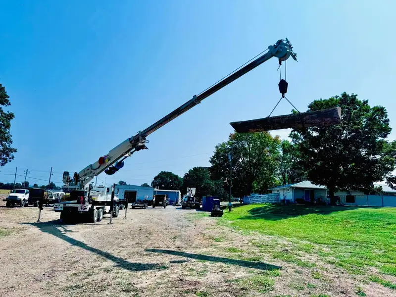 A crane truck lifting a long log high over a grassy lot