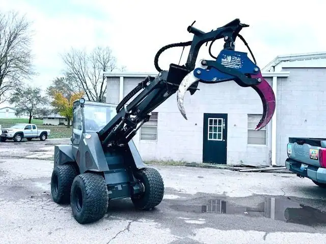 A compact loader with a log grapple attachment parked by a white block building